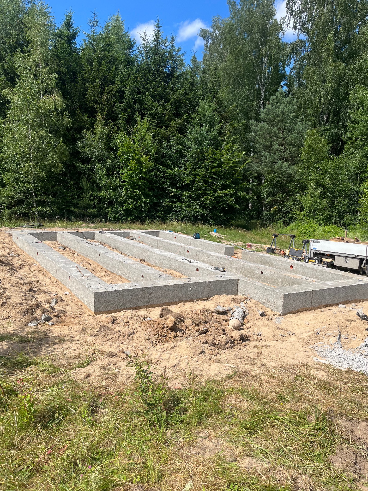 Zalany betonowy szalunek fundamentowy z czerwonymi zaślepkami na tle zieleni i nieba, widoczny przyczepka z narzędziami.