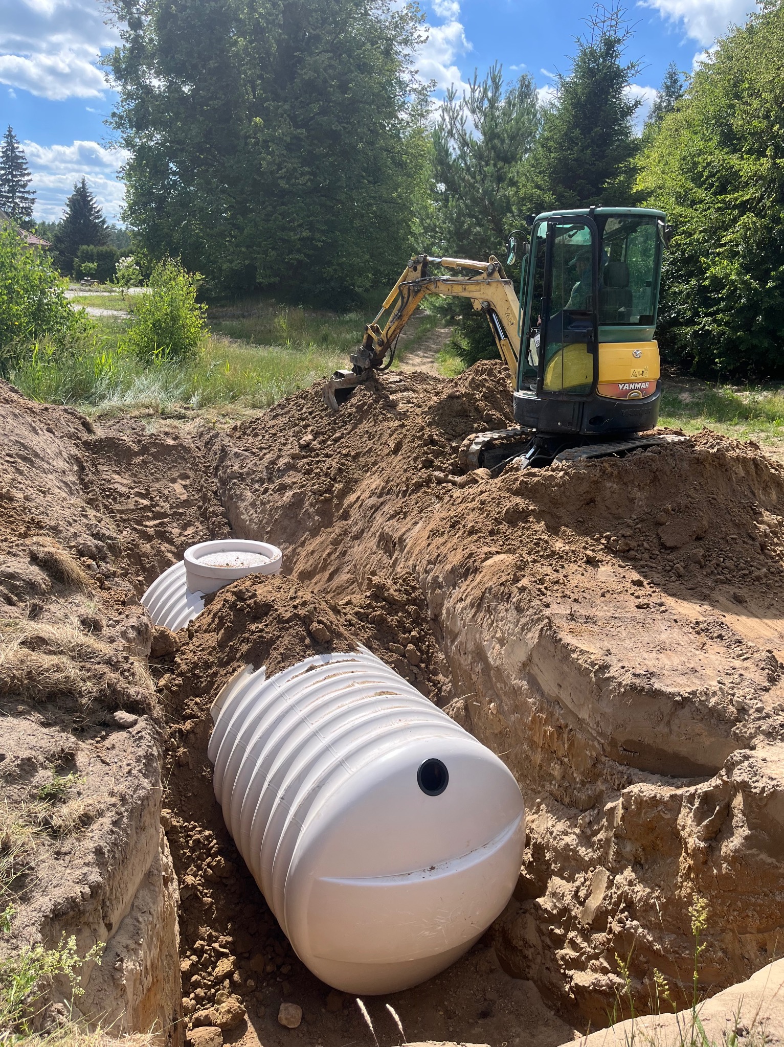 Żółta mini koparka Yanmar pracuje przy wykopie, w którym umieszczono dwa białe zbiorniki retencyjne, widoczne ślady pracy koparki na ziemi.