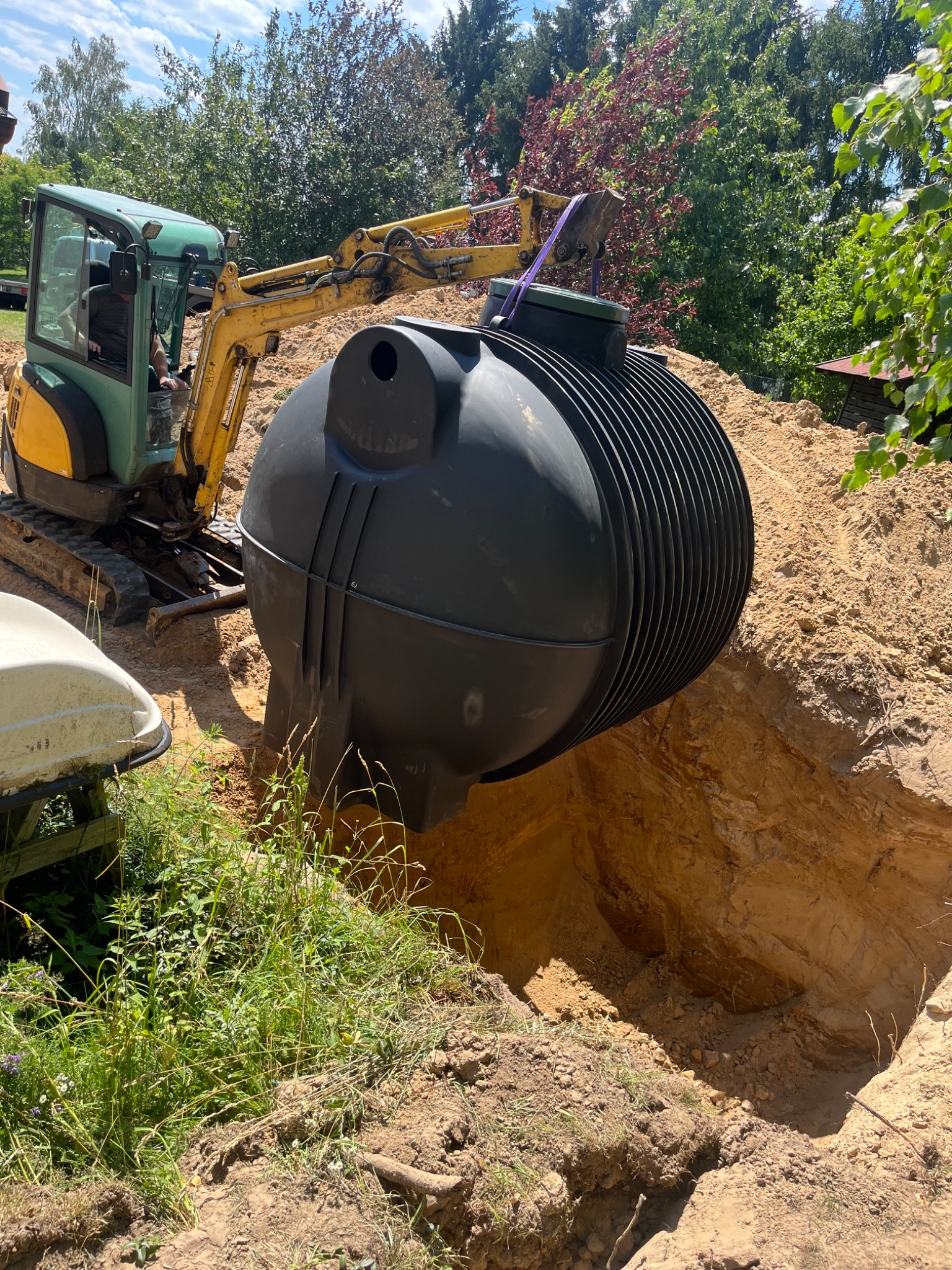 Żółto-zielona mini koparka umieszcza czarny, żebrowany zbiornik w wykopie ziemnym na tle drzew i niebieskiego nieba z chmurami.