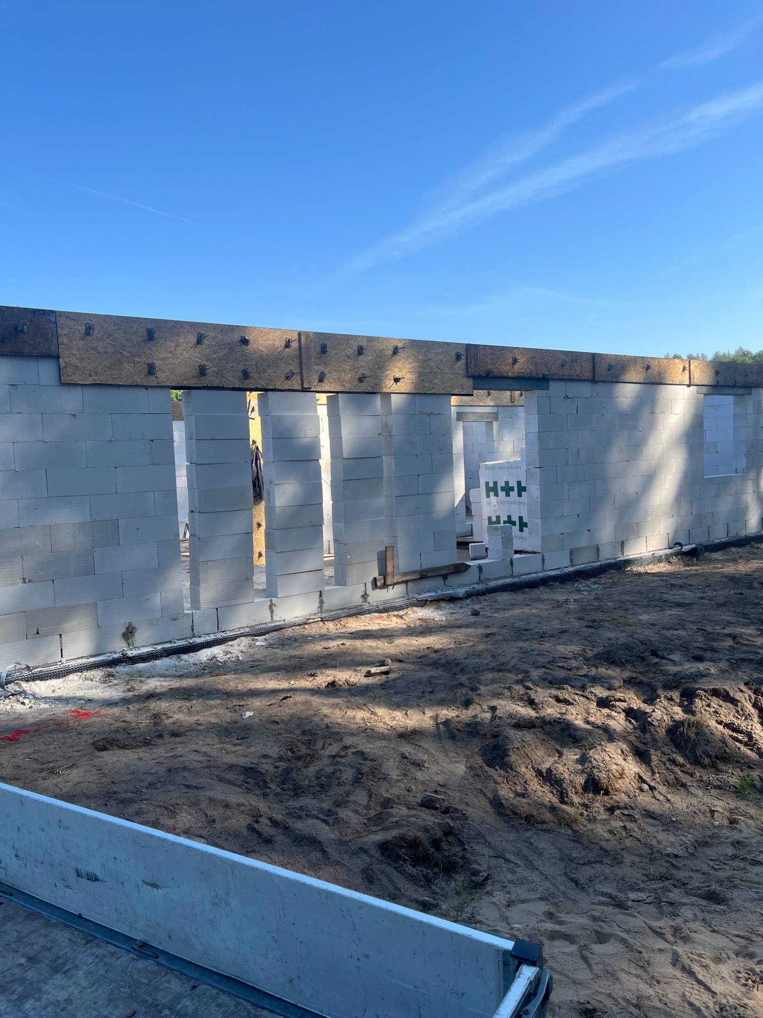 Wznoszone ściany domu z betonu komórkowego H+H, widoczne otwory okienne i drzwiowe, drewniany szalunek górny, teren wokół przygotowany pod dalsze prace budowlane.