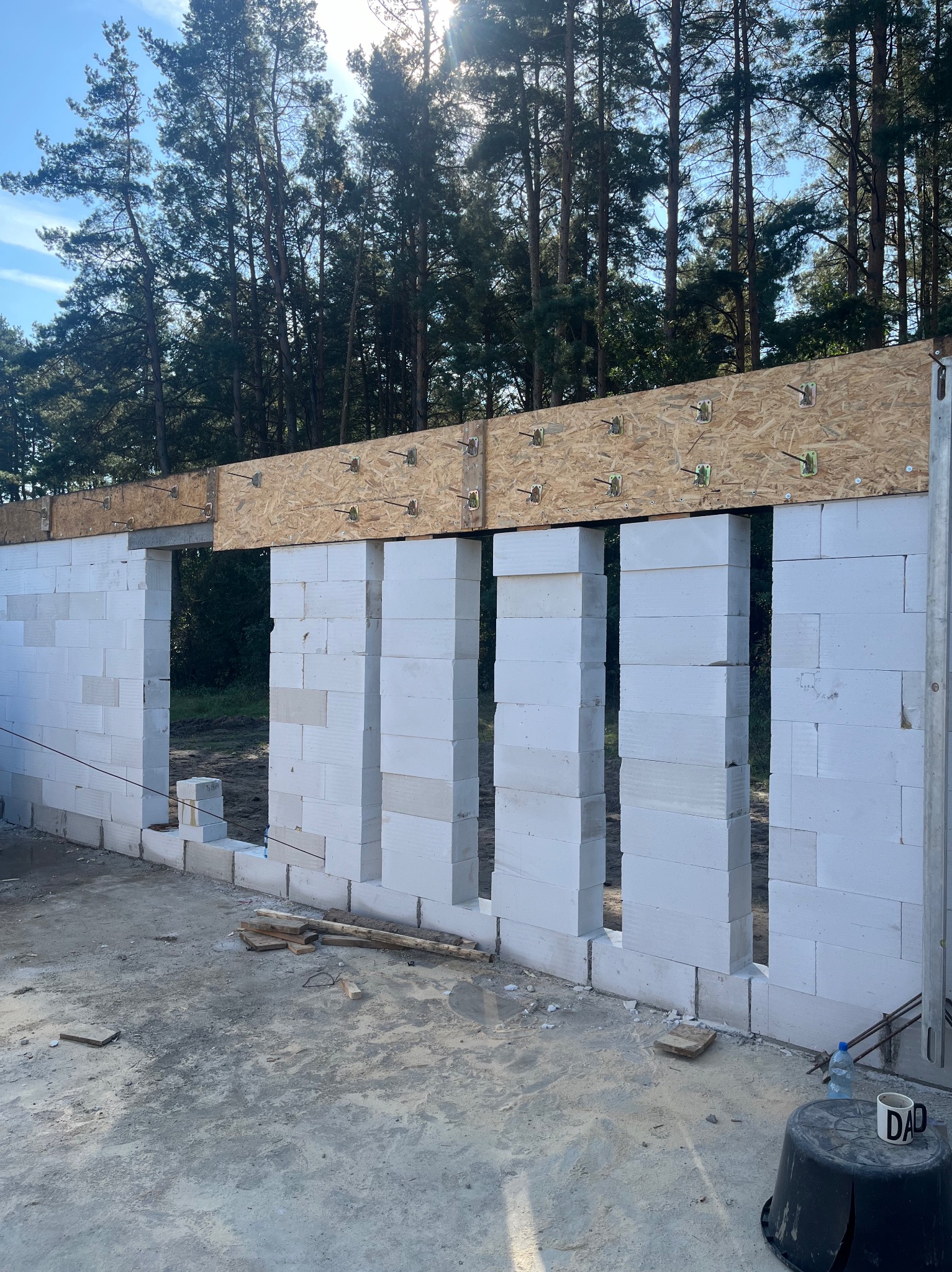 Wznoszenie ścian budynku z białych bloczków, widoczne drewniane elementy konstrukcyjne stropu, w tle las iglasty i błękitne niebo, na pierwszym planie czarne wiadro z napisem 'DAD' i butelka wody.