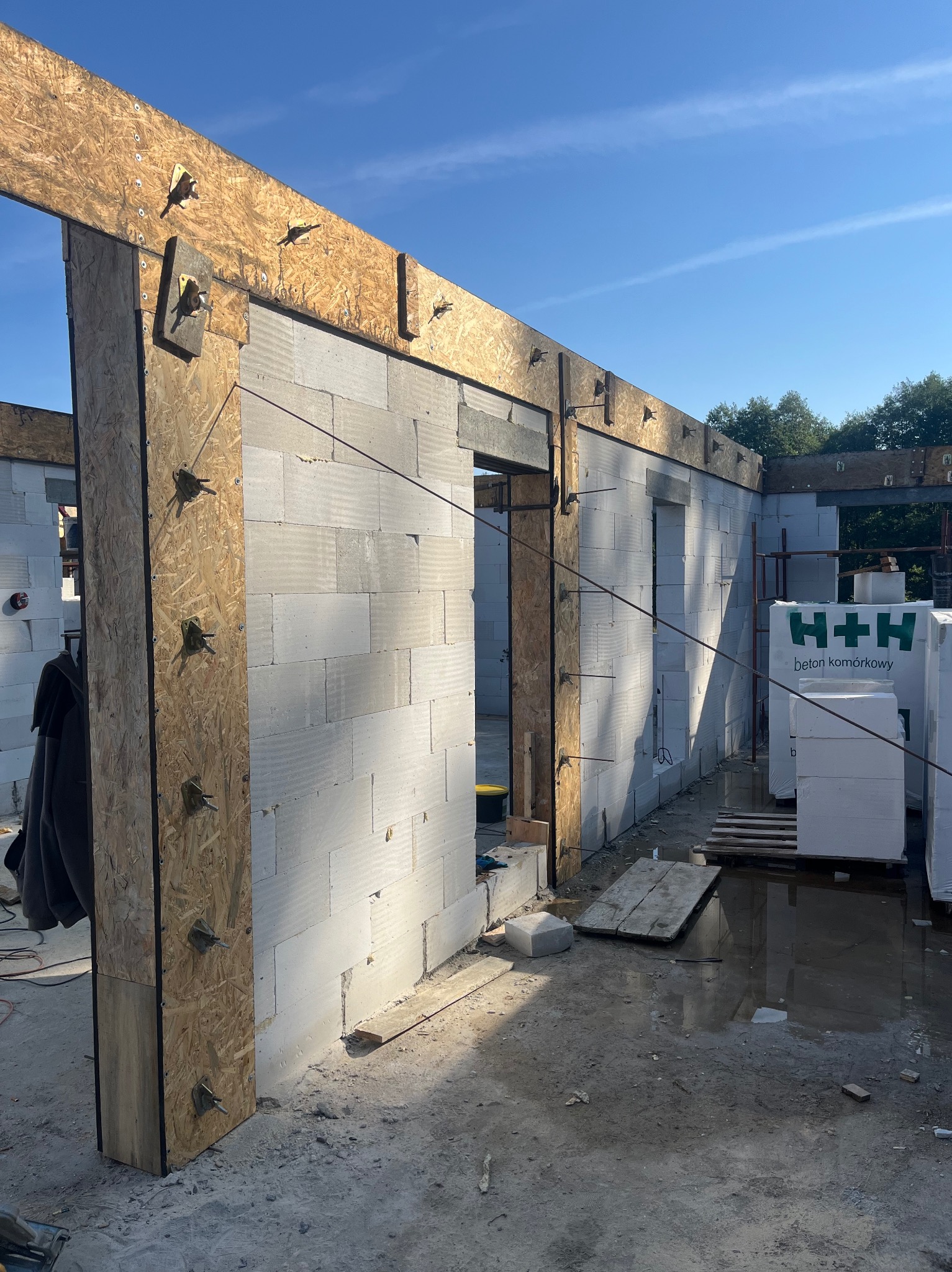 Wznoszenie ścian budynku z betonu komórkowego, widoczne drewniane szalunki, elementy mocujące i palety z bloczkami H+H, teren wokół placu budowy częściowo zalany wodą.