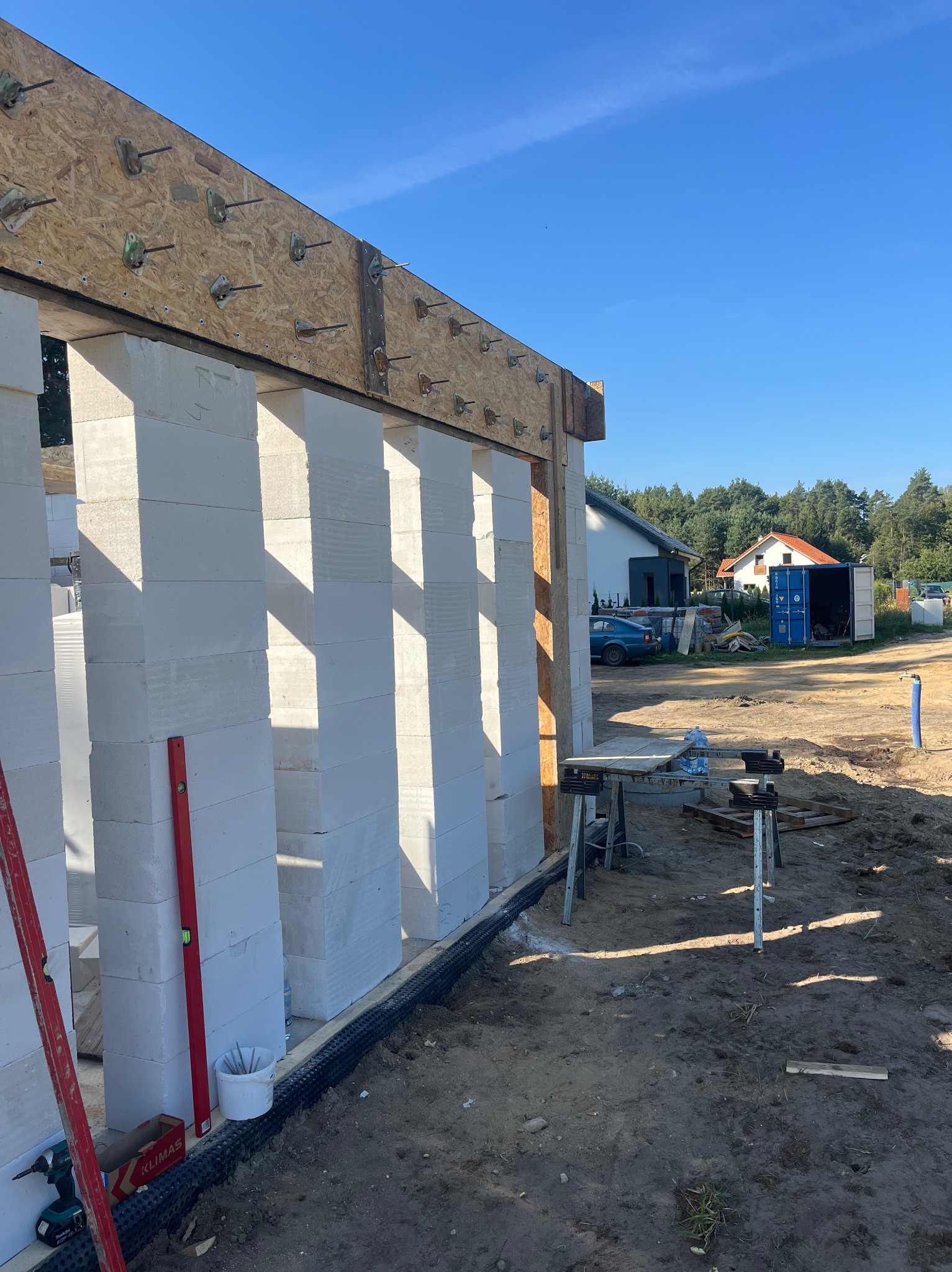 Wznoszenie ścian z bloczków betonowych na fundamencie, widoczne elementy konstrukcyjne, poziomica, narzędzia, oraz otoczenie placu budowy z kontenerem i zabudową jednorodzinną w tle.