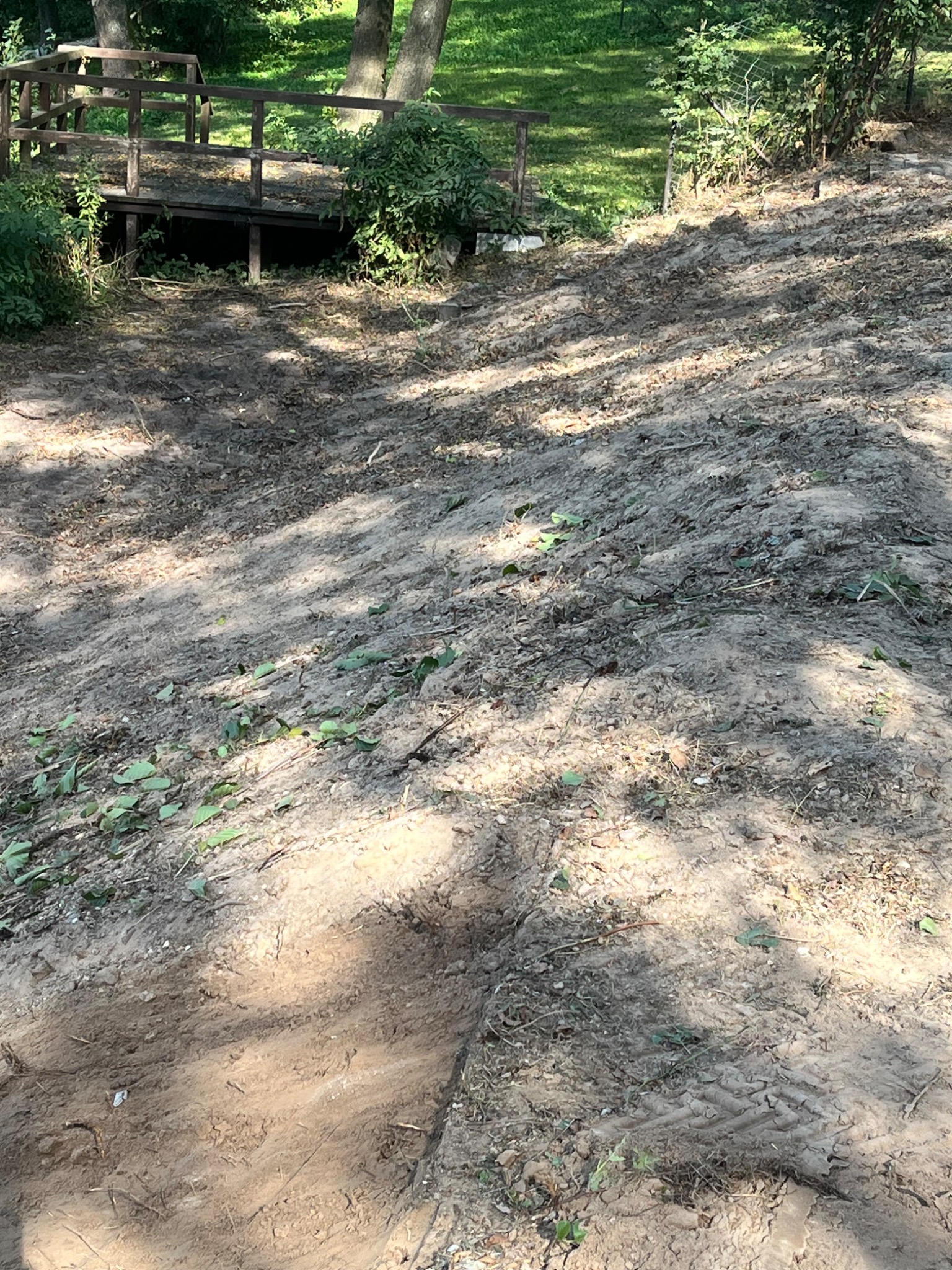 Nierówny teren po niwelacji, z widocznym śladem opon i fragmentem drewnianego mostku w tle.