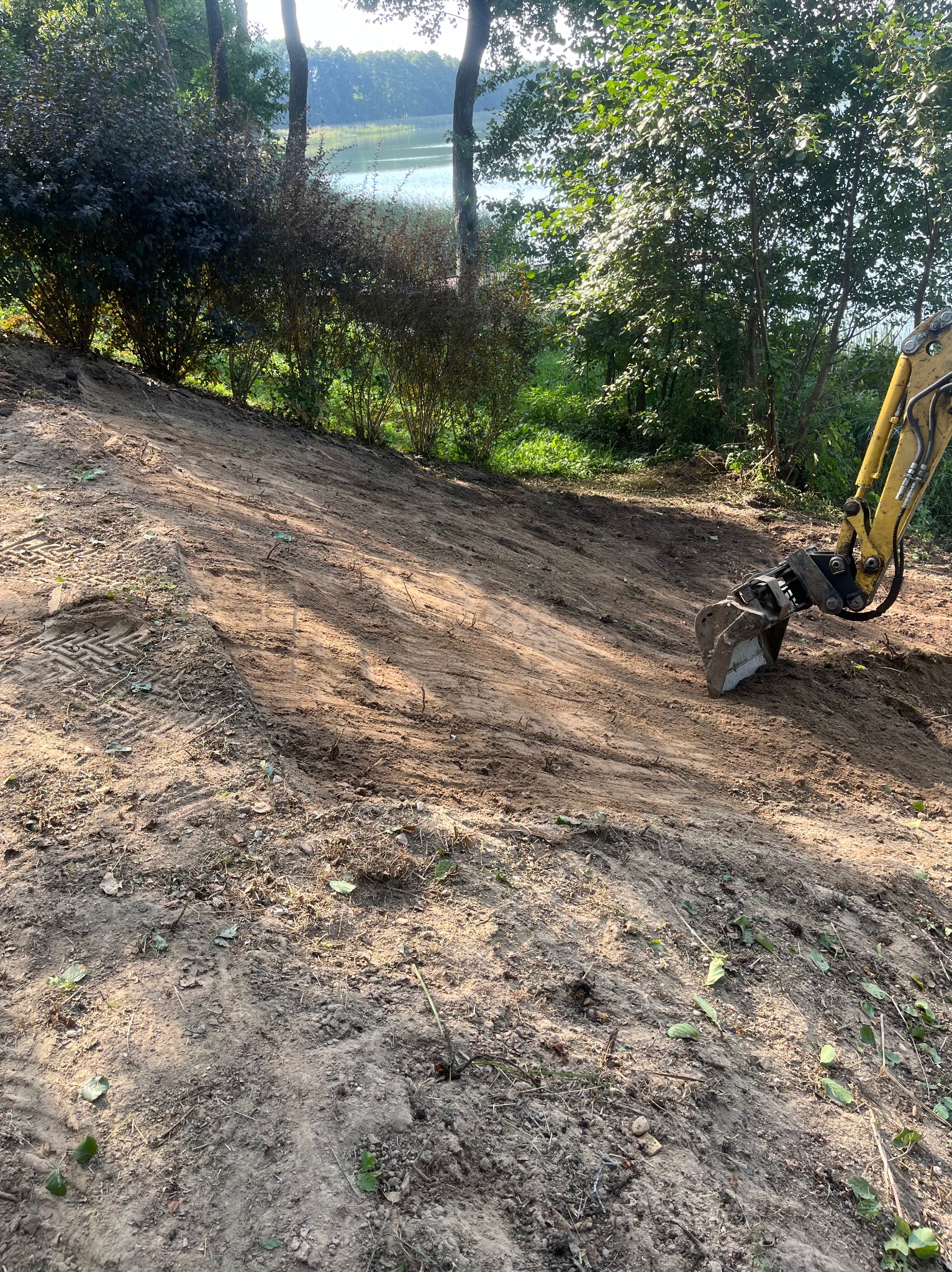 Żółta koparka częściowo widoczna podczas wyrównywania terenu na zboczu w pobliżu drzew i jeziora, widoczne ślady opon.