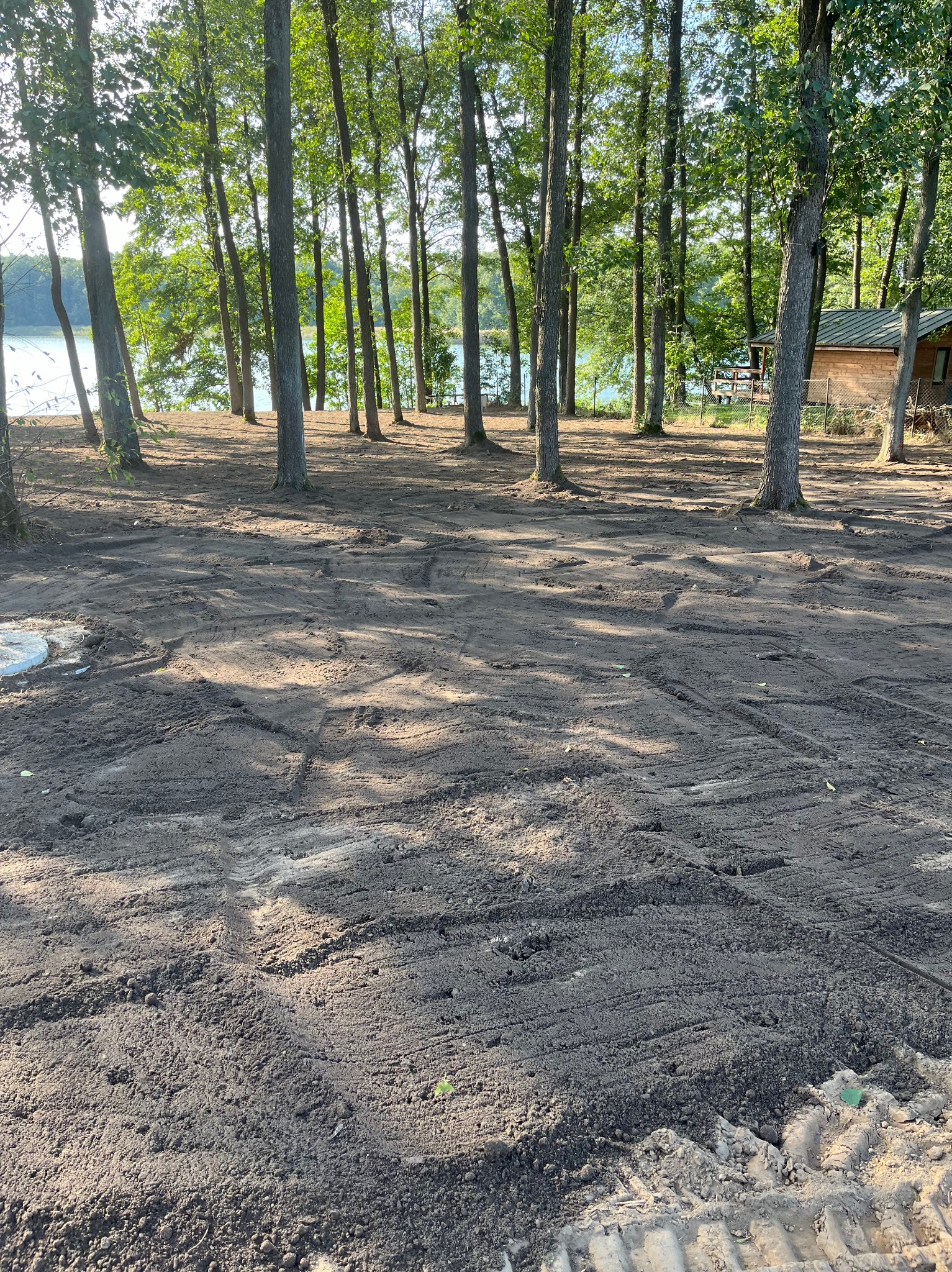 Przygotowany teren pod trawnik, widoczne ślady po maszynach, drzewa w tle, jezioro i domek letniskowy w oddali.
