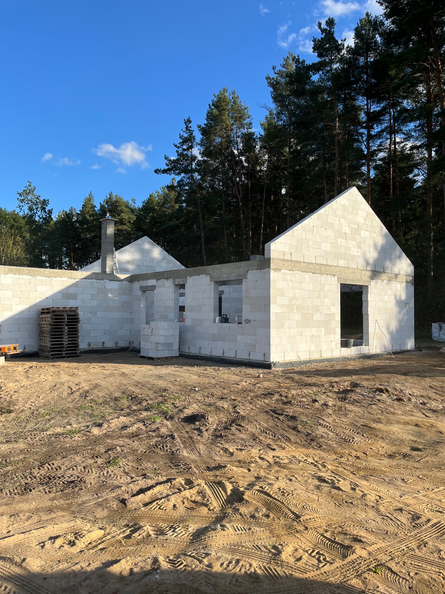 Surowy stan domu jednorodzinnego w trakcie budowy, ściany wzniesione z białych bloczków, widoczne palety i otaczający teren z piaskiem i śladami opon.