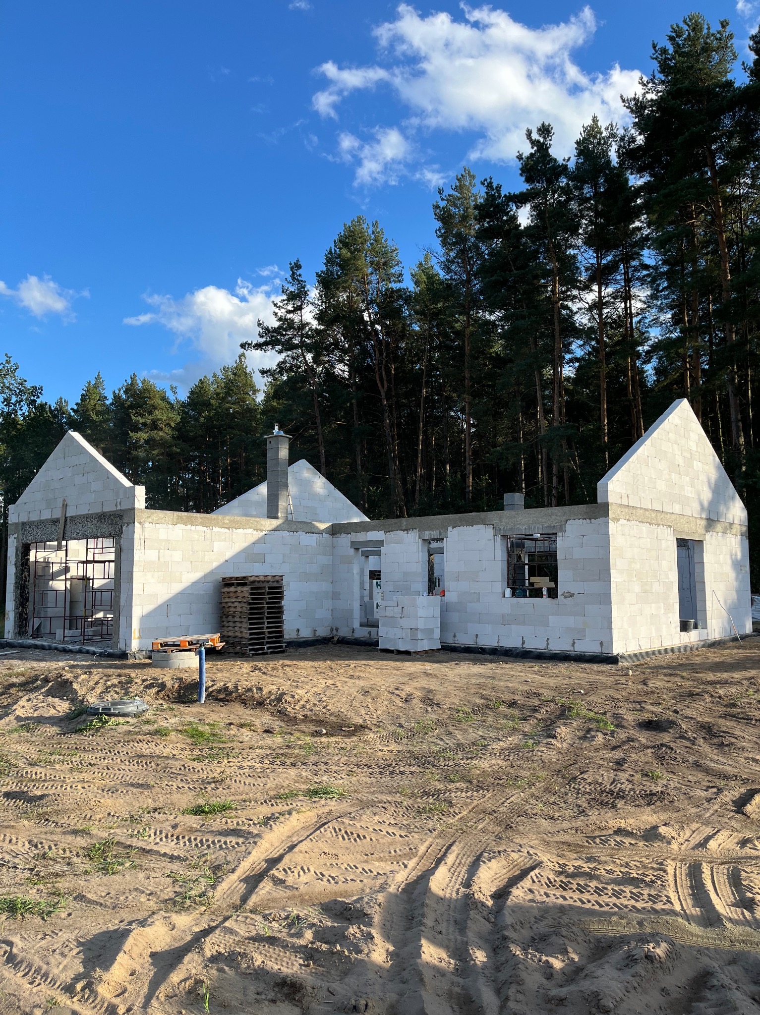 Wznoszenie ścian parterowego domu z białych bloczków, widoczne zbrojenia, paleta i ślady opon na piaszczystym gruncie, w tle wysokie sosny i błękitne niebo.