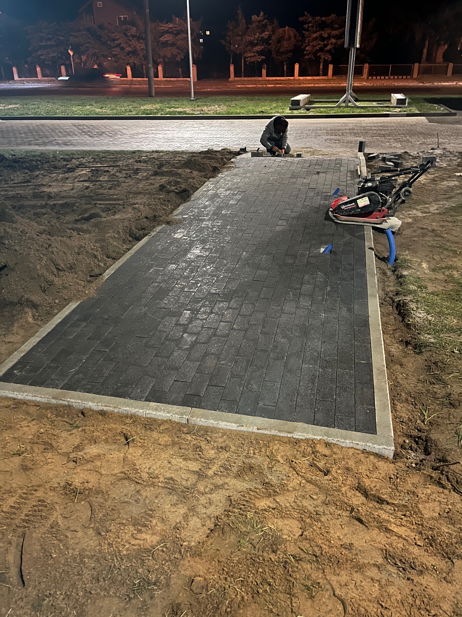 Układanie ciemnej kostki brukowej na posesji, widoczny pracownik i zagęszczarka. Częściowo ułożona nawierzchnia z obrzeżami, przygotowany teren pod dalsze prace.