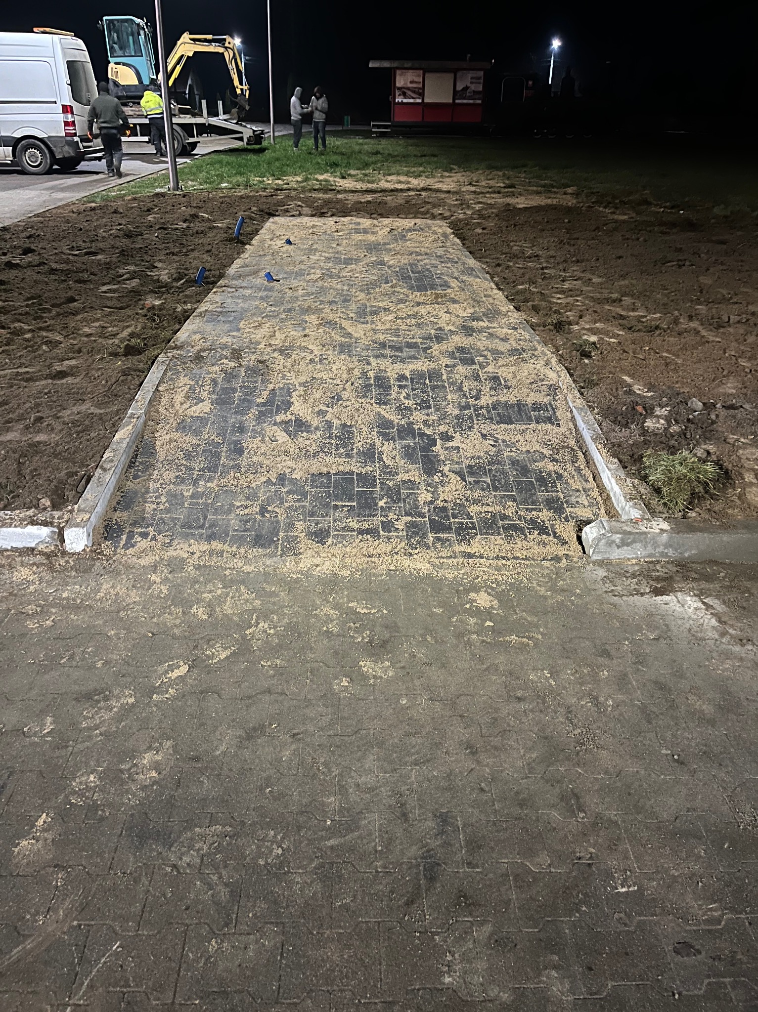 Ułożona kostka brukowa, posypana piaskiem, z obrzeżami. W tle ekipa i koparka na placu budowy nocą. Widoczne ślady pracy.