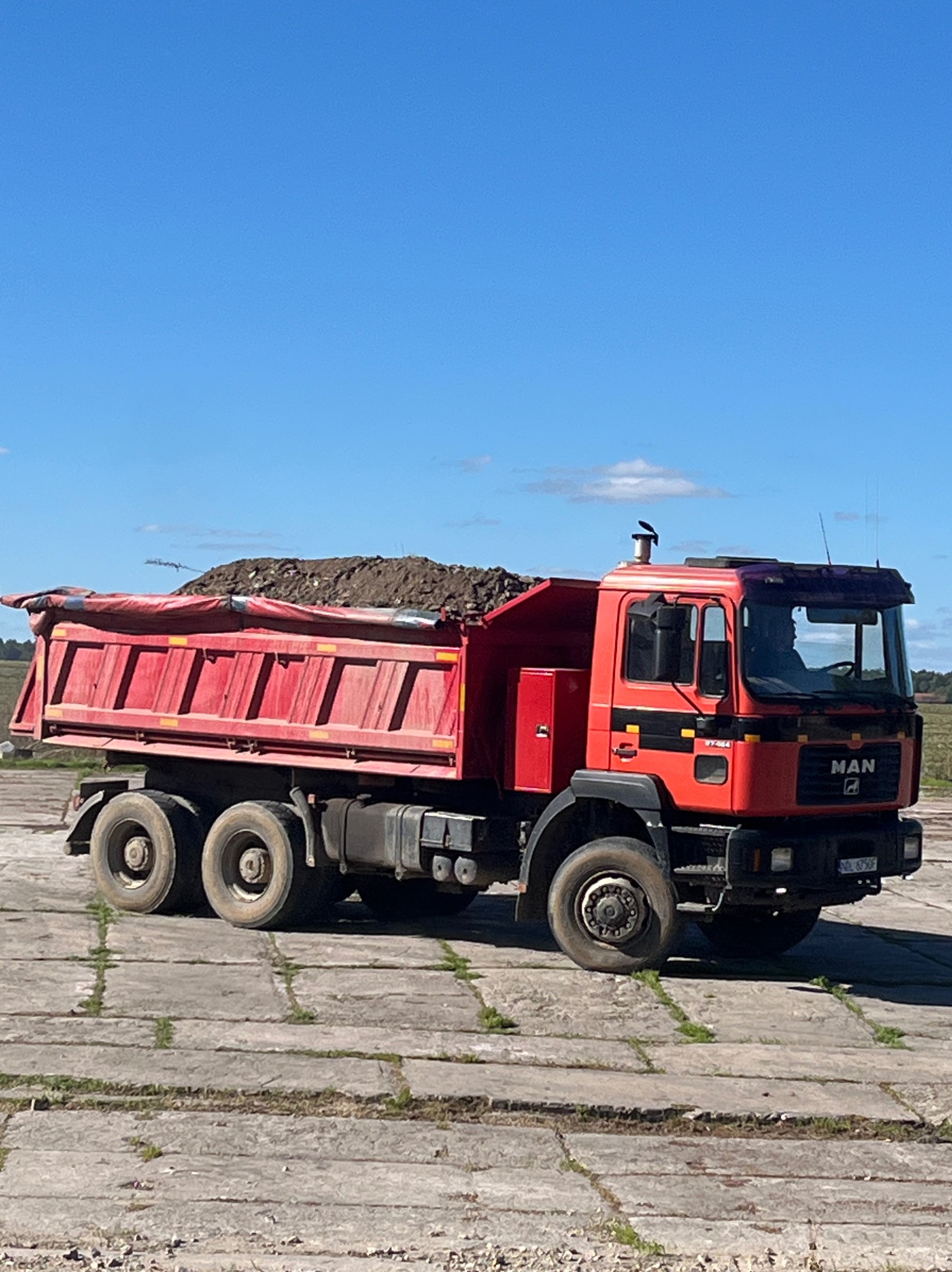 Czerwona ciężarówka MAN z pełną skrzynią ziemi, stojąca na betonowej nawierzchni w słoneczny dzień. Widoczne detale pojazdu i ładunku.