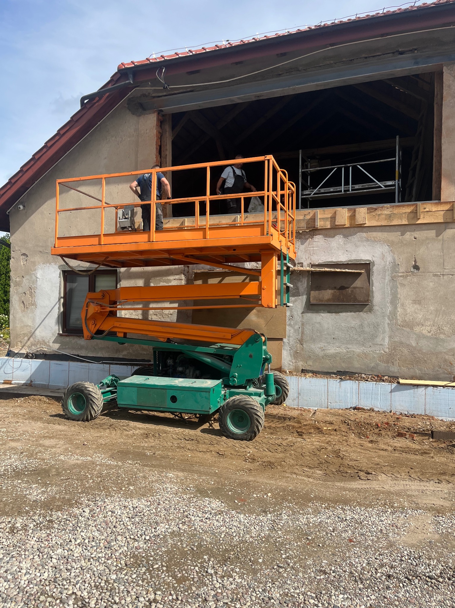 Prace budowlane przy użyciu podnośnika koszowego na tle budynku z otwartym poddaszem. Widoczne przygotowania do dalszych etapów budowy.