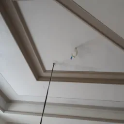 Wykończony sufit z dekoracyjnymi sztukateriami, widoczne kable wystające z otworu oraz zabezpieczone miejsce na oświetlenie, jasna kolorystyka.