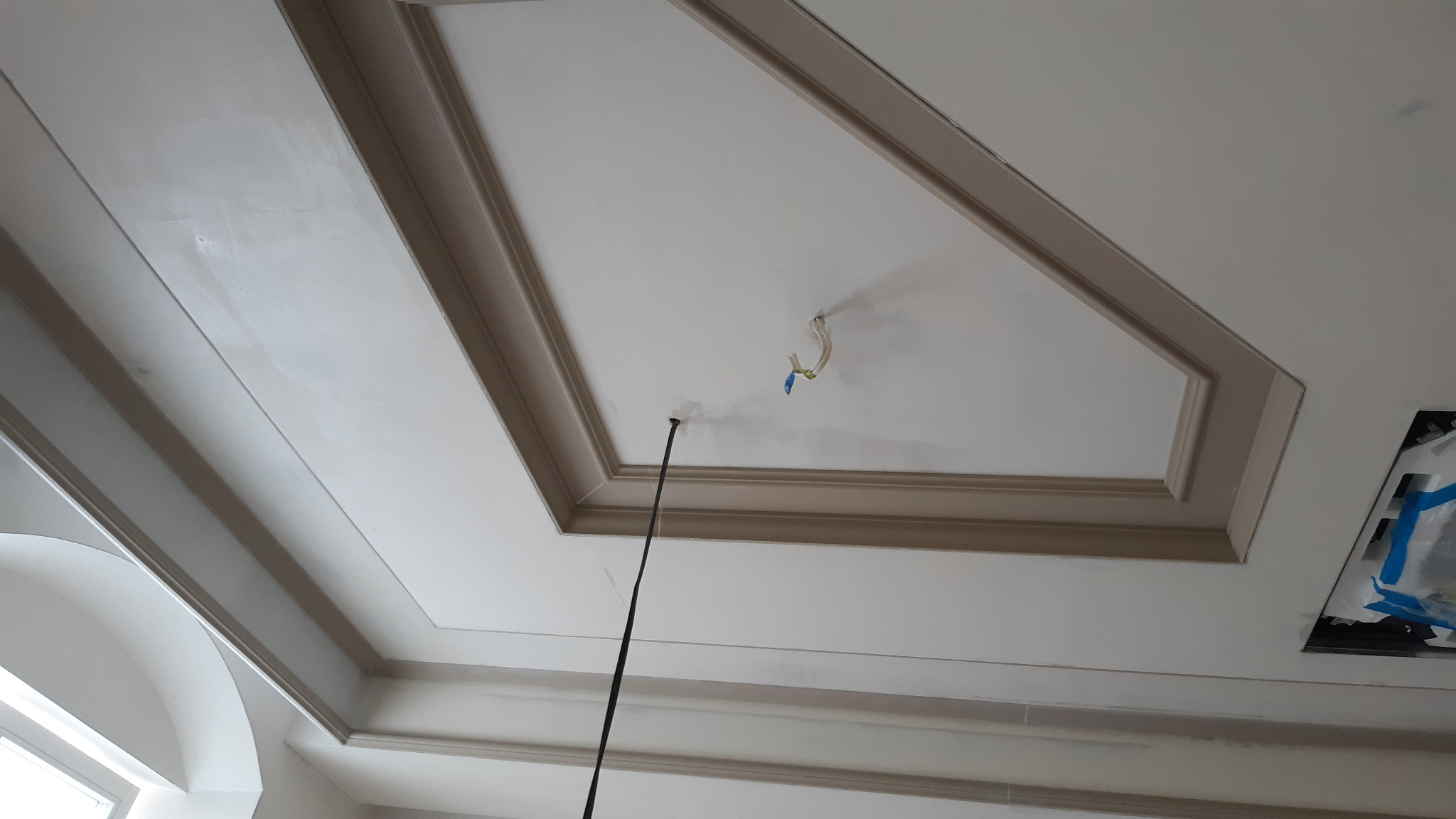 Wykończony sufit z dekoracyjnymi sztukateriami, widoczne kable wystające z otworu oraz zabezpieczone miejsce na oświetlenie, jasna kolorystyka.