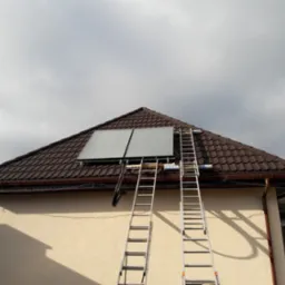 Dwa panele słoneczne zamontowane na skośnym dachu domu z brązową dachówką, podparte dwiema aluminiowymi drabinami, widok z dołu na tle pochmurnego nieba.