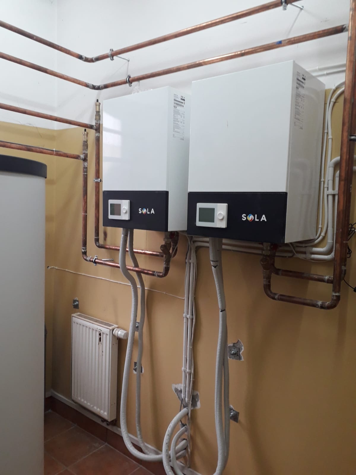 Dwie białe jednostki grzewcze Sola z czarnym panelem, zamontowane na żółtej ścianie z widocznymi miedzianymi rurami i białymi przewodami, obok grzejnika i zbiornika na wodę.