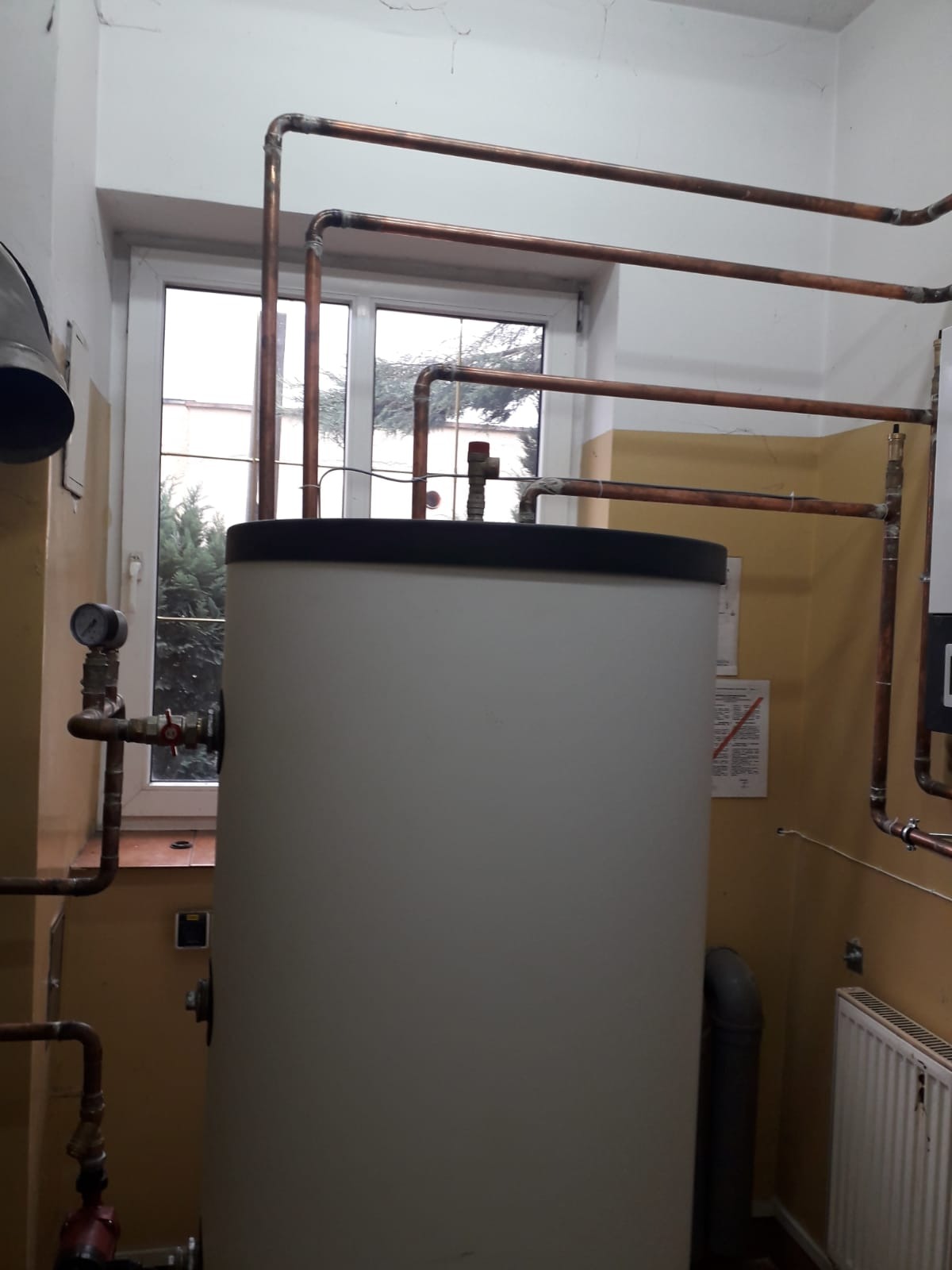 Szary zasobnik ciepłej wody użytkowej z miedzianymi rurami w kotłowni, widoczne okno z widokiem na drzewa i manometr na rurze.