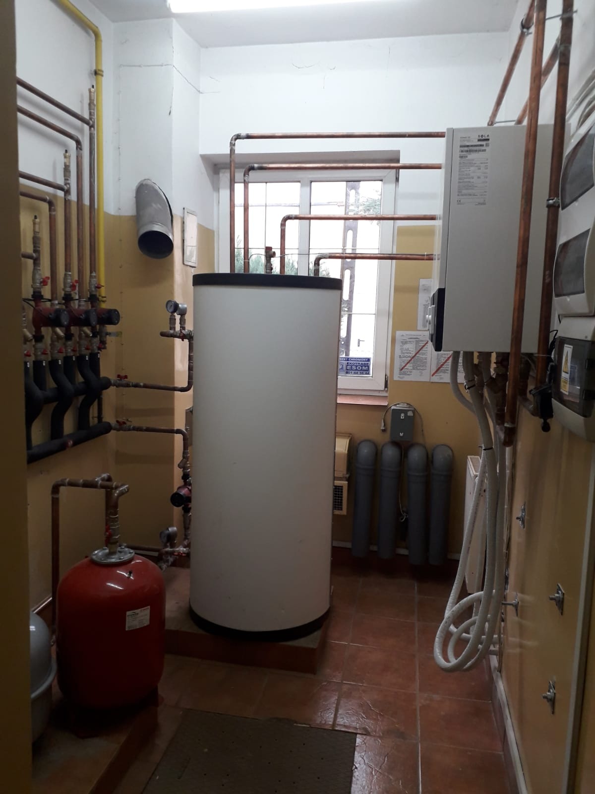 Kotłownia z białym zasobnikiem ciepłej wody, czerwonym naczyniem przeponowym i licznymi miedzianymi rurami w pomieszczeniu z terakotą na podłodze.