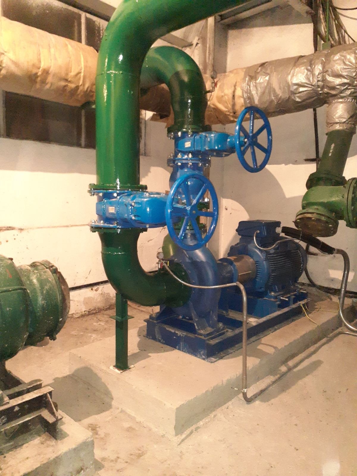 Zestaw rur w kolorze zielonym i niebieskim z zaworami i pompą na betonowej podstawie w piwnicy.