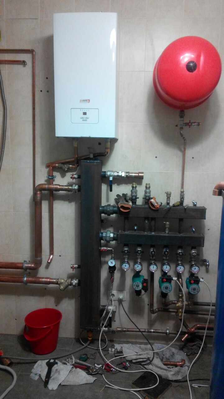 Nowa instalacja grzewcza z białym piecem, czerwonym zbiornikiem wyrównawczym i rozdzielaczem, widoczne miedziane rury, manometry, pompy obiegowe oraz narzędzia hydrauliczne na podłodze.