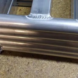 Fragment spawanej konstrukcji z rur i profili aluminiowych, widoczne spoiny i przygotowane otwory montażowe, leżącej na płycie wiórowej.