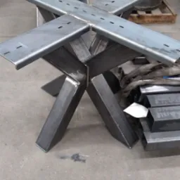 Metalowa podstawa stołu o geometrycznym kształcie krzyża z przyspawanymi płaskownikami z otworami, widoczna w warsztacie obróbki metali, obok leżą profile stalowe.