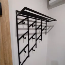 Czarny, metalowy wieszak na ubrania z półką na czapki, o geometrycznej konstrukcji, zamontowany na białej ścianie obok drewnianej framugi drzwi.