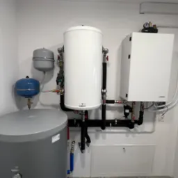 Zestaw urządzeń grzewczych w kotłowni: biały zbiornik akumulacyjny Galmet, szary zasobnik, biały kocioł Hewalex z osprzętem, rury w izolacji, manometry i zawory, całość zamontowana na ścianie...