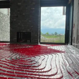Czerwone rury ogrzewania podłogowego ułożone na srebrnej folii z siatką w surowym wnętrzu domu z kamiennym kominkiem i dużym oknem z widokiem na krajobraz.