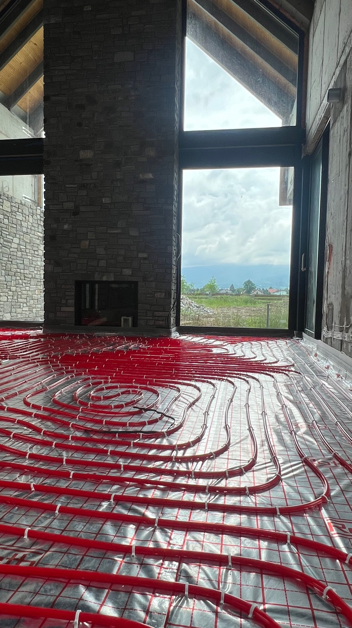 Czerwone rury ogrzewania podłogowego ułożone na srebrnej folii z siatką w surowym wnętrzu domu z kamiennym kominkiem i dużym oknem z widokiem na krajobraz.