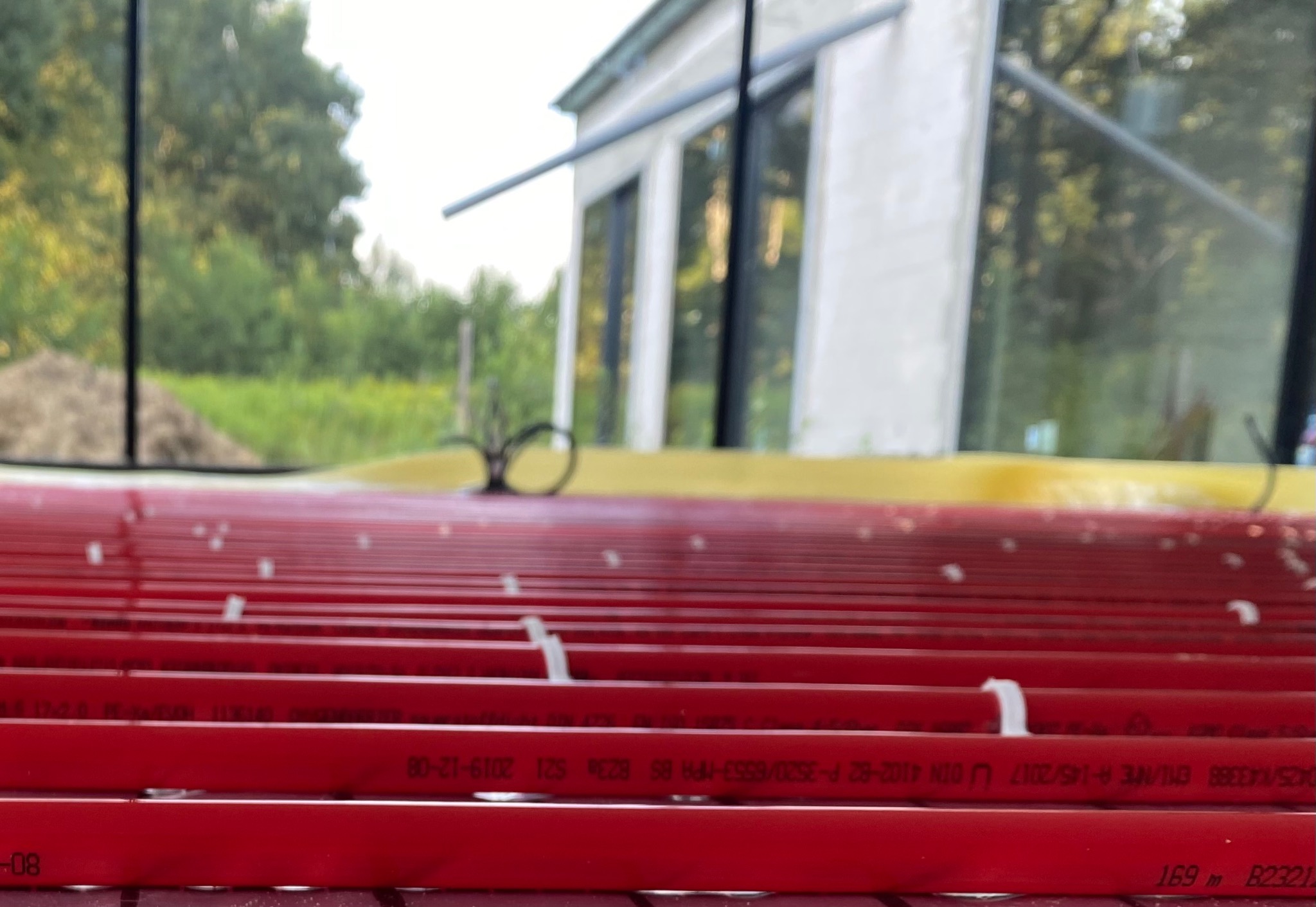 Czerwone rury ogrzewania podłogowego rozłożone na żółtej izolacji, widoczne napisy na rurach, w tle okna budynku i zieleń