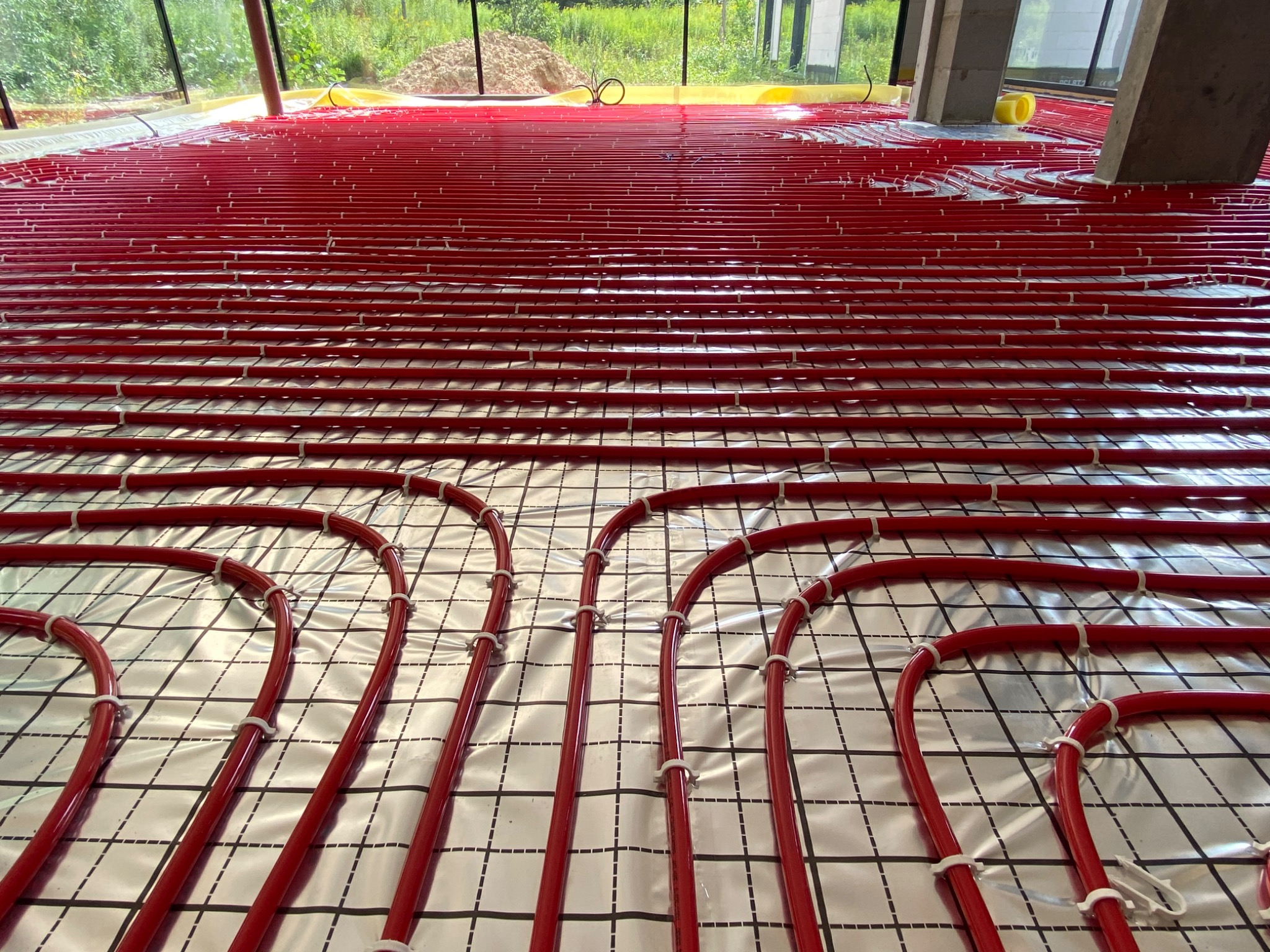 Czerwone rury ogrzewania podłogowego ułożone na srebrnej folii z rastrem, zabezpieczone białymi uchwytami, widok z bliska na zakręty rur.