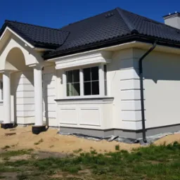 Nowoczesny dom jednorodzinny z jasną elewacją, ciemną dachówką i dekoracyjnymi elementami architektonicznymi, widoczne przygotowania do zagospodarowania terenu wokół budynku.