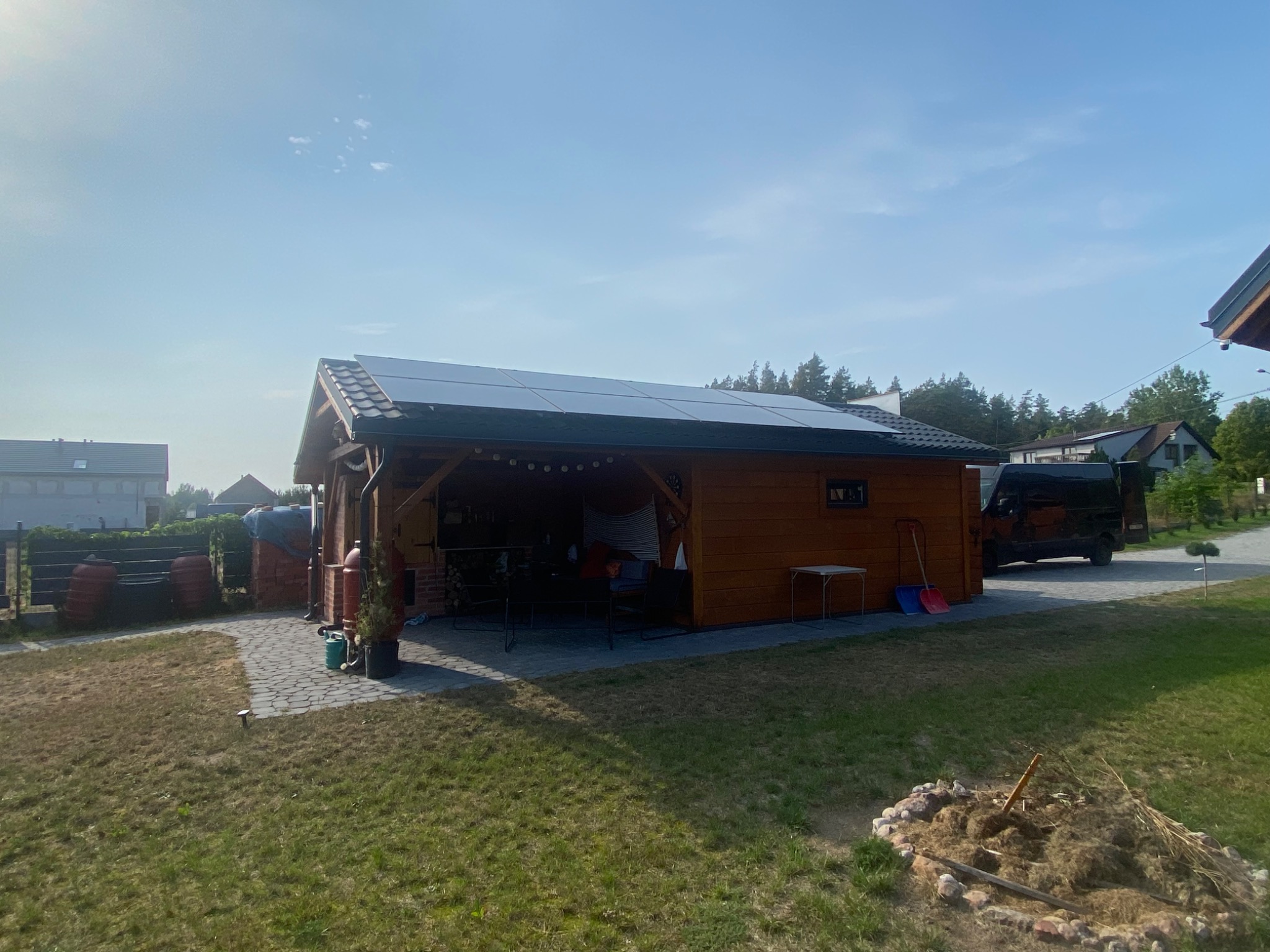 Drewniana altana z panelami słonecznymi na dachu, oświetlona girlandą żarówek, widoczna na tle zielonego trawnika i błękitnego nieba.