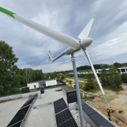 Widok z góry na turbinę wiatrową z zielonymi akcentami na końcach łopat, zamontowaną na dachu budynku z panelami fotowoltaicznymi, w tle zieleń i zabudowa jednorodzinna.