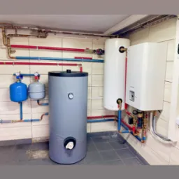 Instalacja centralnego ogrzewania w kotłowni z szarym zbiornikiem akumulacyjnym, dwoma niebieskimi naczyniami wzbiorczymi, białym bojlerem i wiszącym kotłem gazowym, połączonymi rurami miedzianymi...
