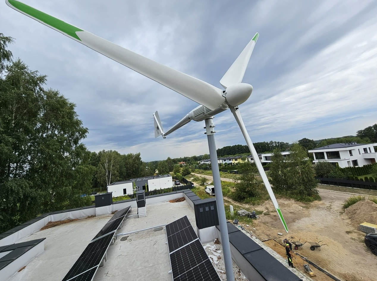 Widok z góry na turbinę wiatrową z zielonymi akcentami na końcach łopat, zamontowaną na dachu budynku z panelami fotowoltaicznymi, w tle zieleń i zabudowa jednorodzinna.