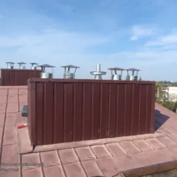 Brązowy komin z metalowymi nasadami na dachu z blachy w odcieniach brązu, widok z góry na tle błękitnego nieba.