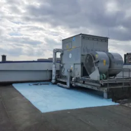 Płaski dach pokryty świeżą, jasnoniebieską warstwą hydroizolacyjną, z widoczną instalacją wentylacyjną i fragmentami innych budynków w tle.