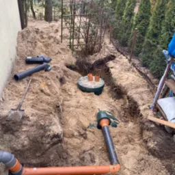 Montaż instalacji sanitarnej w wykopie ziemnym: rury kanalizacyjne, studzienka rewizyjna z pomarańczowymi rurami. W tle iglaki i drabina.