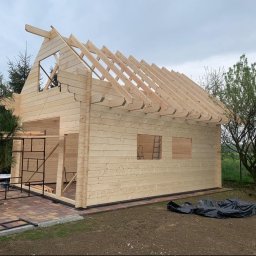 Uslugi Budowlane Minikop Zwierzak Przemyslaw Graczyk - Surowy drewniany szkielet domu w trakcie budowy. Widoczne krokwie dachowe, ściany z bali i rusztowania. Budynek otoczony zielenią, w tle pochmurne niebo.
