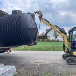 Uslugi Budowlane Minikop Zwierzak Przemyslaw Graczyk - Żółta koparka Yanmar ostrożnie zdejmuje czarny zbiornik z naczepy ciężarówki na tle domów i zieleni. Widoczne pakiety materiałów izolacyjnych.