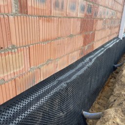 Uslugi Budowlane Minikop Zwierzak Przemyslaw Graczyk - Ocieplenie fundamentów budynku z bloczków ceramicznych, zabezpieczone folią kubełkową i drenażem. Widok na fragment ściany i wykop z instalacją.