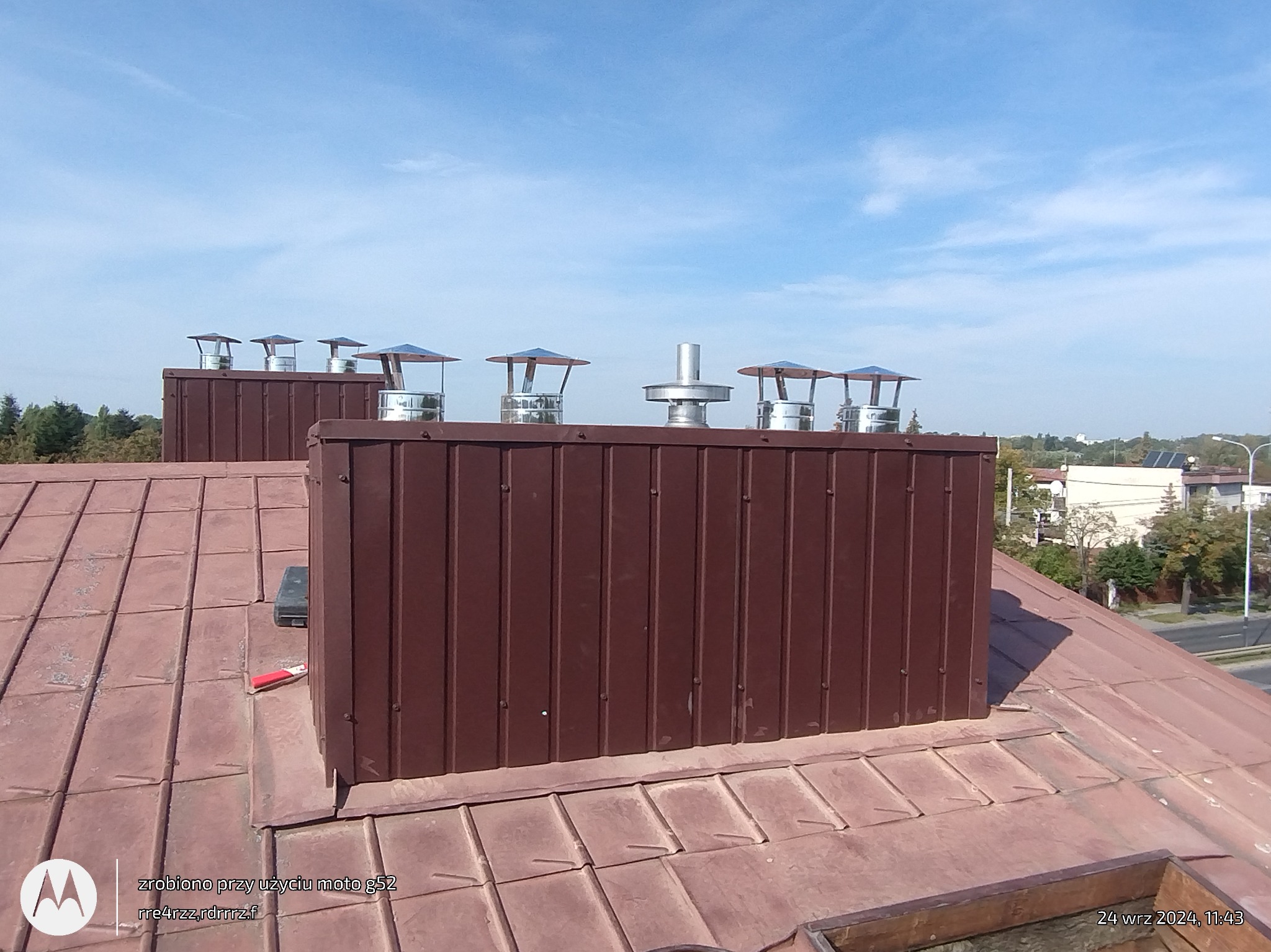 Brązowy komin z metalowymi nasadami na dachu z blachy w odcieniach brązu, widok z góry na tle błękitnego nieba.