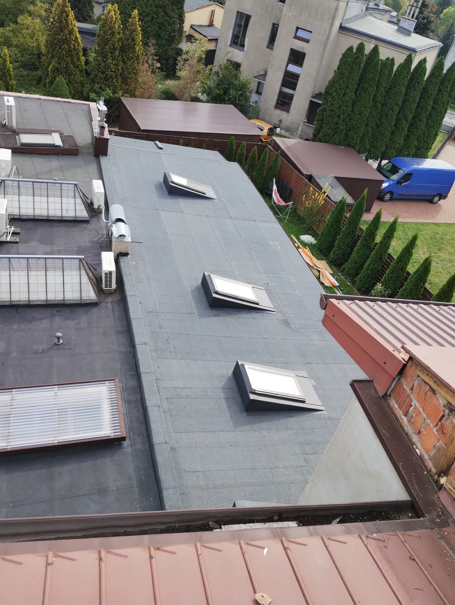 Widok z góry na płaski dach pokryty papą z trzema oknami dachowymi i klimatyzatorami, w tle budynki mieszkalne i zieleń, widoczny fragment dachu z blachy trapezowej i rynną.