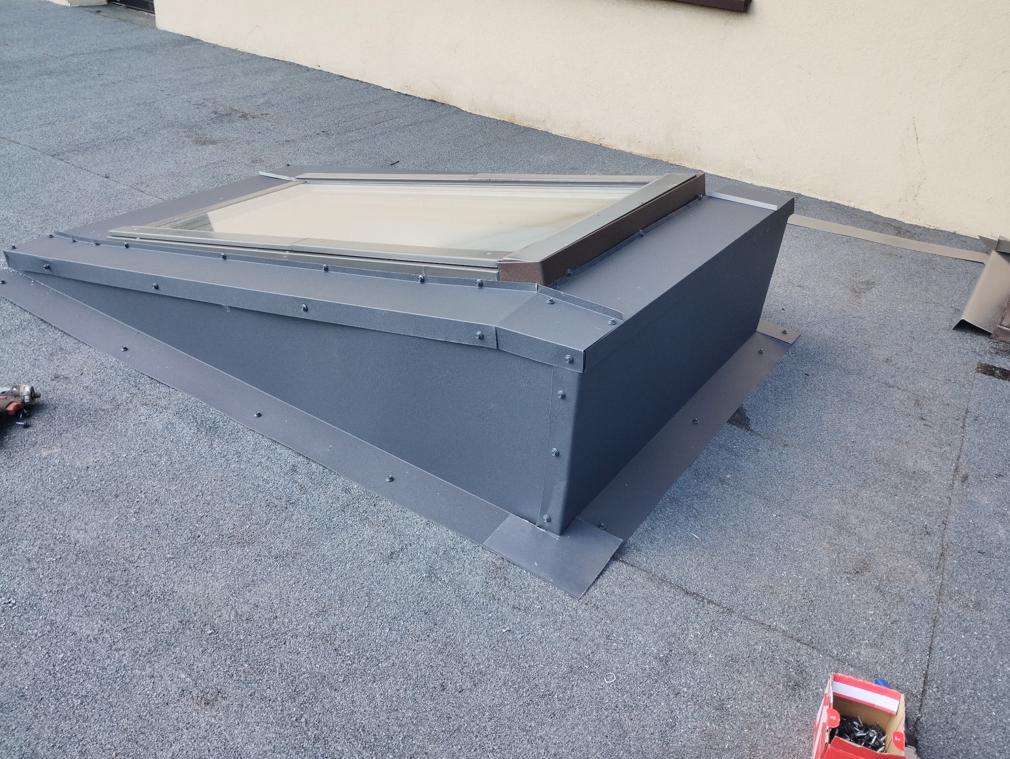 Szare okno dachowe z brązowymi elementami wykończeniowymi zamontowane na pokryciu dachowym z papy, widoczne narzędzia i akcesoria montażowe.