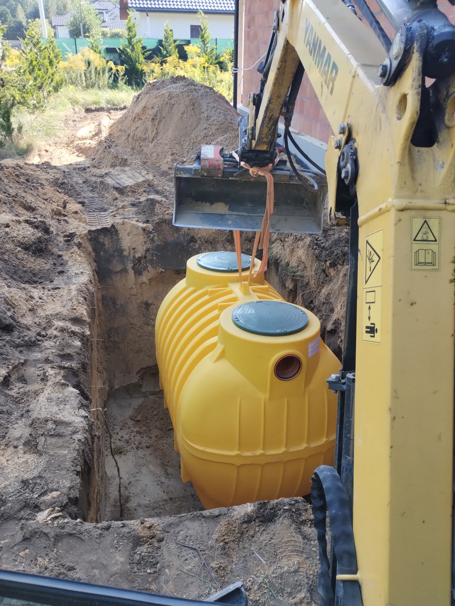 Żółty zbiornik z dwoma zielonymi pokrywami, podwieszony na pasach w łyżce koparki Yanmar, umieszczany w wykopie ziemnym. Na ziemi widoczne ślady opon koparki i hałda wykopanej ziemi. W tle dom...