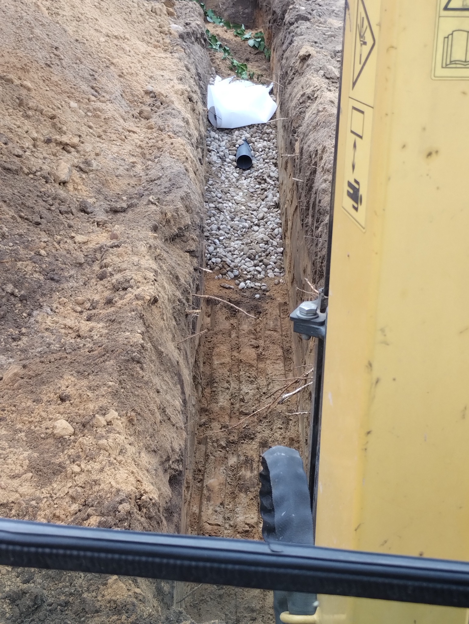 Wykopany wąski rów z widocznym drenażem z rurą i geowłókniną, widziany z kabiny żółtej koparki.