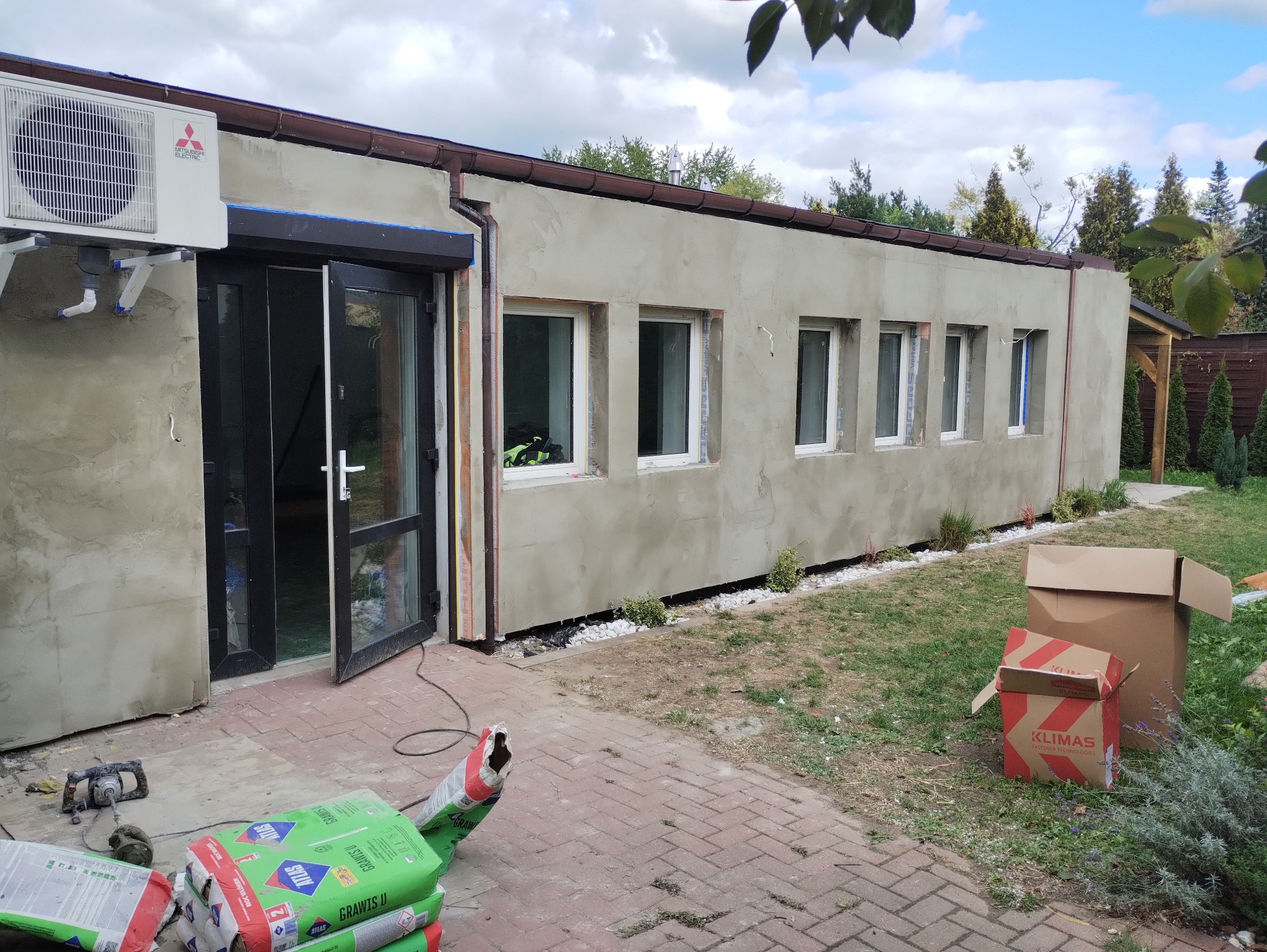 Budynek po nałożeniu szarego tynku zewnętrznego, z zamontowanymi oknami PCV i otwartymi czarnymi drzwiami, widoczny klimatyzator, worki z zaprawą i narzędzia na brukowanym podłożu, teren budowy.