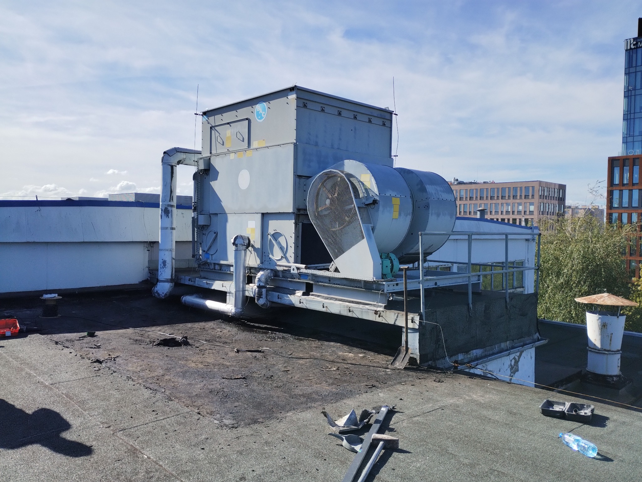 Szary system wentylacji przemysłowej z dwoma wentylatorami na płaskim dachu, z widocznymi elementami instalacji i narzędziami, w tle budynki miejskie i błękitne niebo.